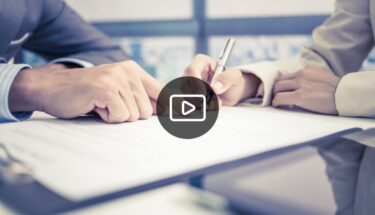 An image of two people's hands writing on a document, there is a play button to suggest this is for a video.