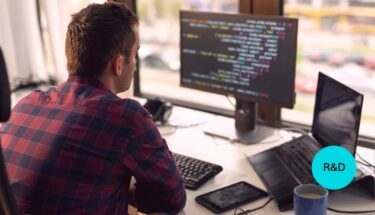R&D professional sitting on a desk and looking at a computer screen with code.