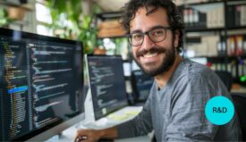A smiling man with glasses sitting on a desk with a computer