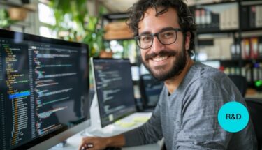 A smiling man with glasses sitting on a desk with a computer