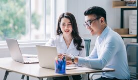 A woman and a man professionally dressed sitting down and looking at computer screens while smiling