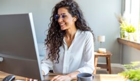 A woman with white shirt and glasses smiling while typing on her computer