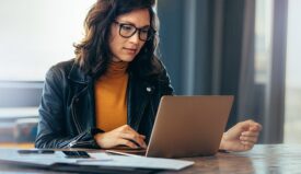 A woman wearing a leather jacket and glasses sitting down and looking at her computer screen