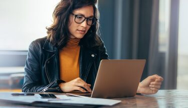 A woman wearing a leather jacket and glasses sitting down and looking at her computer screen
