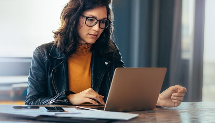 A woman wearing a leather jacket and glasses sitting down and looking at her computer screen