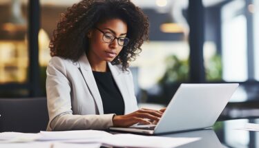 A professionally dressed woman sitting down on a desk and looking into a computer