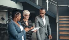 A man and two women walking and talking while looking at a tablet