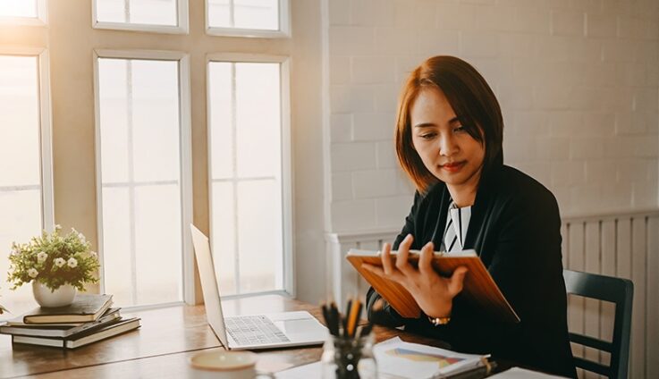 A woman reading a document in a desk next to computer