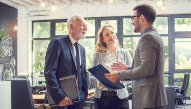 Three professionally dressed people in an office setting speaking with each other