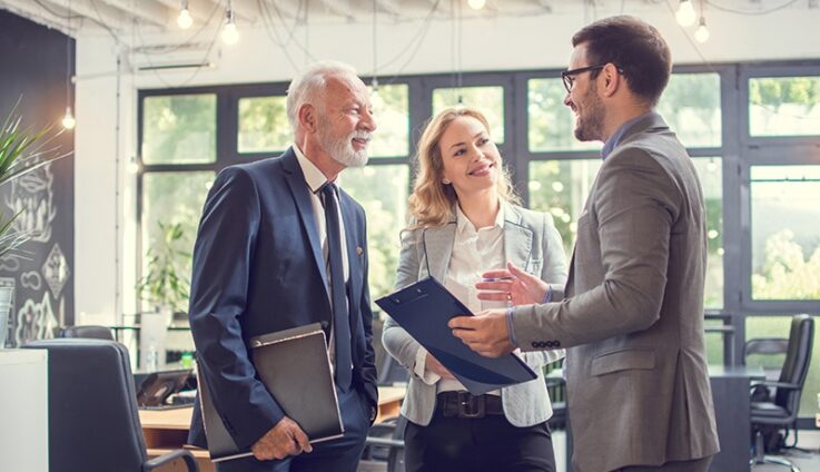 Three professionally dressed people in an office setting speaking with each other