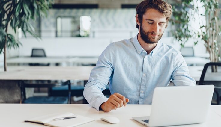A white man in a light blue button down shirt sits at a table with a white laptop open in front of him.