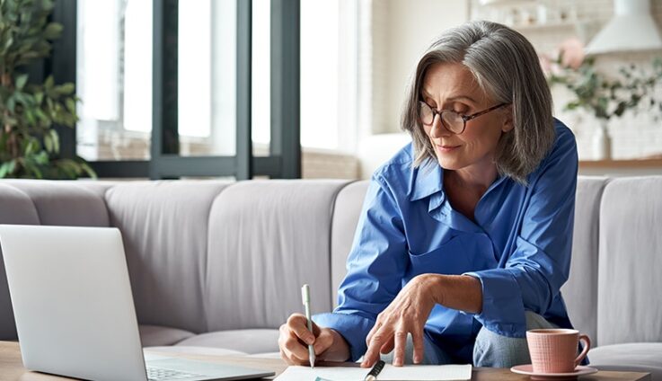 A woman with glasses taking notes on a sheet of paper while sitting on a desk