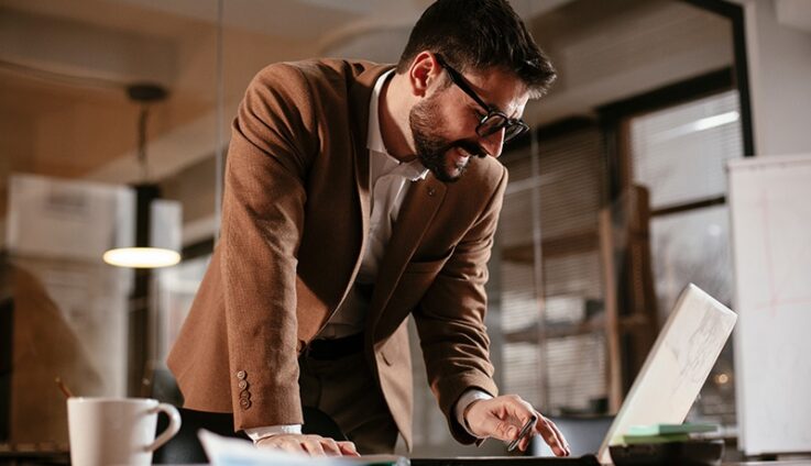 Professionally dressed man with glasses smiling and looking at a computer.