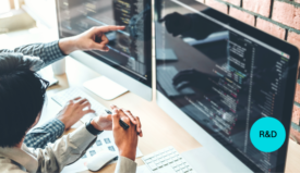 We see two sets of hands in front of two monitor screens at a desk. One hand is pointing to something on a monitor.