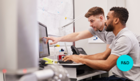 Two men work at a computer. One is typing, the other is pointing to something on the monitor.