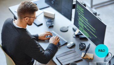 A white man with glasses is coding at a computer at a black desk.