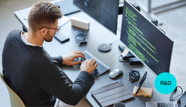 A white man with glasses is coding at a computer at a black desk.