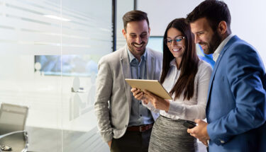 A group of young professionals in business attire smiling and looking at a tablet.