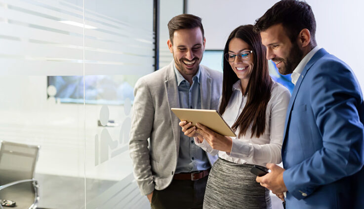 A group of young professionals in business attire smiling and looking at a tablet.
