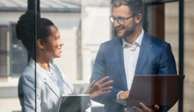 A man and women, both dressed professional, hold tablets and talk.