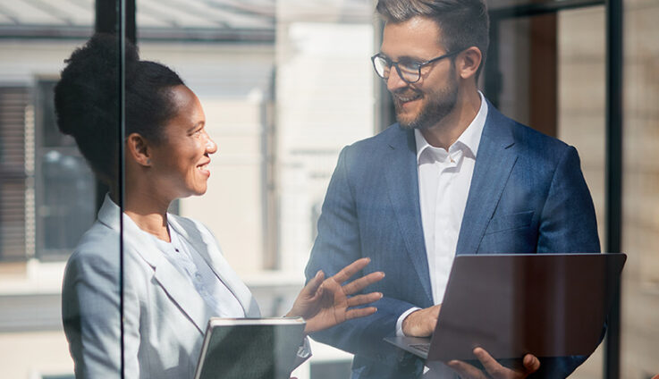 A man and women, both dressed professional, hold tablets and talk.