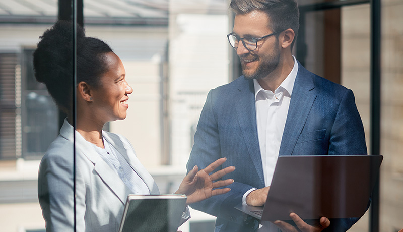 A man and women, both dressed professional, hold tablets and talk.